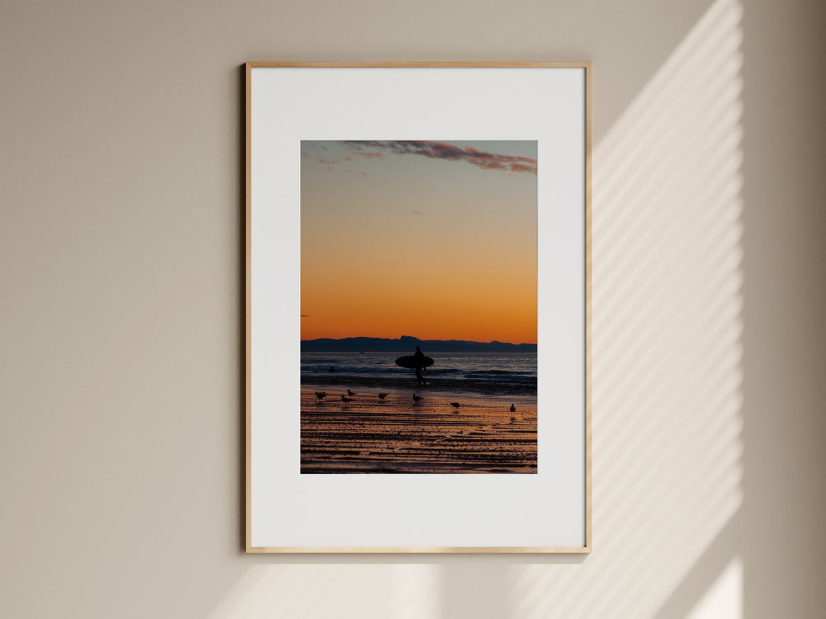 Natural oak framed surf photography print of a surfer at sunset hanging on a beige living room wall