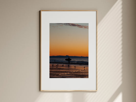 Natural oak framed surf photography print of a surfer at sunset hanging on a beige living room wall