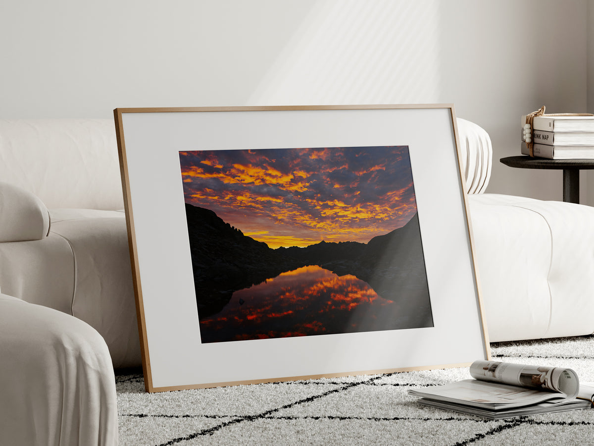 Natural oak framed mountain landscape print of a fiery sunrise reflection standing on the floor in a bright modern room