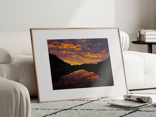 Natural oak framed mountain landscape print of a fiery sunrise reflection standing on the floor in a bright modern room